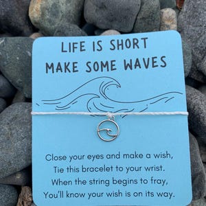 May include: A light blue card with the text "LIFE IS SHORT MAKE SOME WAVES" and a wave illustration. A silver-coloured charm bracelet with a wave design hangs from the card. The card also includes instructions.