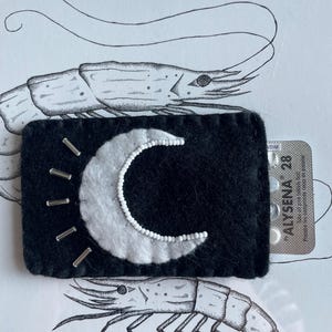 Peut inclure: Un rectangle de feutre noir avec un croissant de lune blanc orné de petites perles blanches. Des épingles argentées rayonnent de la lune. L'image comprend un blister de pilules et un dessin de crevette.