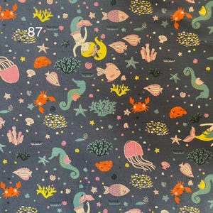 Pode incluir: Tecido azul escuro com um padr&atilde;o repetido de criaturas marinhas. O design inclui sereias, peixes, cavalos-marinhos, caranguejos, &aacute;guas-vivas, corais e estrelas-do-mar em tons de laranja, rosa, amarelo e verde-azulado. O n&uacute;mero "87" &eacute; vis&iacute;vel no canto superior esquerdo.