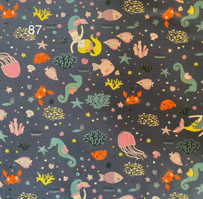 Pode incluir: Tecido azul escuro com um padr&atilde;o repetido de criaturas marinhas. O design inclui sereias, peixes, cavalos-marinhos, caranguejos, &aacute;guas-vivas, corais e estrelas-do-mar em tons de laranja, rosa, amarelo e verde-azulado. O n&uacute;mero "87" &eacute; vis&iacute;vel no canto superior esquerdo.