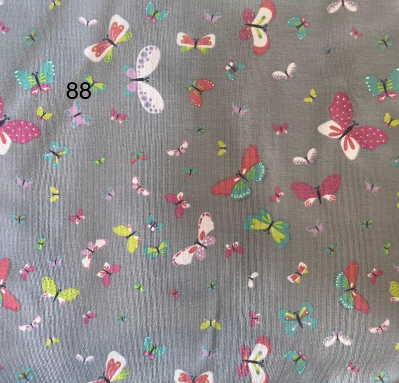 Pode incluir: Tecido cinza coberto por um padr&atilde;o de borboletas coloridas. As borboletas s&atilde;o de v&aacute;rios tamanhos e cores, incluindo rosa, branco, verde e amarelo. O n&uacute;mero "88" est&aacute; impresso no tecido.