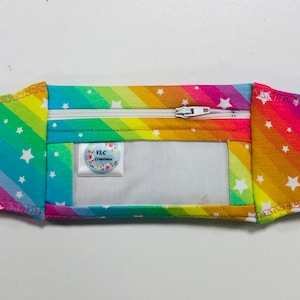 Pode incluir: Uma carteira de tecido com as cores do arco-&iacute;ris, com z&iacute;per branco e janela transparente. A carteira &eacute; decorada com estrelas brancas e tem uma etiqueta com a inscri&ccedil;&atilde;o "VLC Creations".