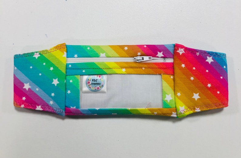 Pode incluir: Uma carteira de tecido com as cores do arco-&iacute;ris, com z&iacute;per branco e janela transparente. A carteira &eacute; decorada com estrelas brancas e tem uma etiqueta com a inscri&ccedil;&atilde;o "VLC Creations".