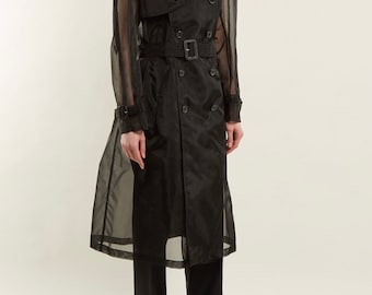 May include: A black trench coat with a sheer, see-through design. The coat features a double-breasted front, a belted waist, and epaulettes on the shoulders. The model is also wearing black pants and black leather shoes.