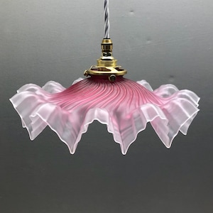 Może przedstawiać: Lampa wisząca z kloszem z matowego, różowego szkła o falistym, marszczonym brzegu. Klosz posiada radialny, teksturowany wzór i jest przymocowany do metalowej oprawki w kolorze mosiądzu oraz szarego przewodu elektrycznego w oplocie z tkaniny.