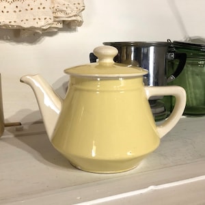 May include: A pale yellow ceramic teapot with a white lid and handle. The teapot has a rounded body and a slightly flared spout.