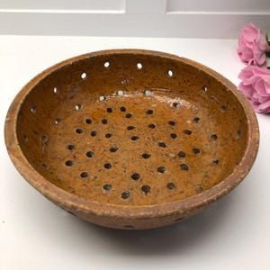 Könnte beinhalten: Eine runde, braune Keramik-Gemüseschüssel mit zahlreichen kleinen Löchern. Die Gemüseschüssel hat eine leicht raue Textur und einen warmen, erdigen Farbton. Der Rand ist ein dunklerer Braunton. Die Gemüseschüssel wird wahrscheinlich zum Ablassen von Flüssigkeiten aus Lebensmitteln verwendet.