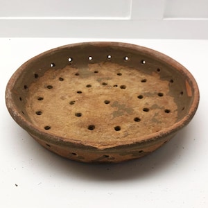 May include: A shallow, circular terracotta bowl with a weathered, earthy brown surface. The bowl features a series of small, evenly spaced drainage holes punched through the base and along the curved sides, showing a porous, unglazed texture.