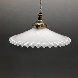 Może przedstawiać: Biała szklana lampa wisząca z szerokim, rozkloszowanym kloszem o falistych, karbowanych krawędziach. Klosz jest przymocowany do oprawki i elementów z polerowanego mosiądzu, zawieszony na skręconym szaro-białym przewodzie elektrycznym w oplocie.