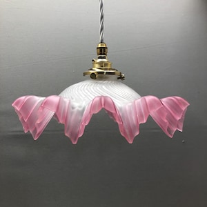 Może przedstawiać: Lampa wisząca z kloszem z matowego szkła o wirującej fakturze i falistym, różowym brzegu. Klosz jest przymocowany do oprawy z polerowanego mosiądzu i wisi na białym przewodzie elektrycznym pokrytym tkaniną.