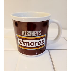 May include: A white ceramic mug with a brown exterior featuring the Hershey's s'mores logo. The mug has a white interior and a curved handle. The mug is on a white tiled surface.