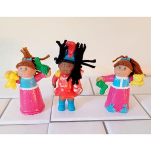 May include: Three vintage Cabbage Patch Kids dolls. Two dolls wear pink dresses with blue accents and hold a yellow bear and a green shoe. The third doll wears a red uniform with a black hat and holds a green shoe.
