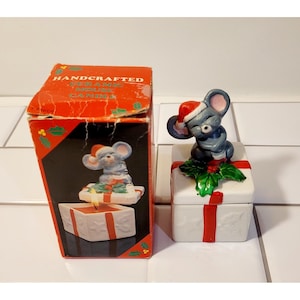 May include: A ceramic mouse figurine wearing a Santa hat sits atop a white gift box with a red ribbon. The box is next to a red box with the text "HANDCRAFTED CERAMIC MOUSE CANDLE". The mouse figurine is gray and has pink ears.