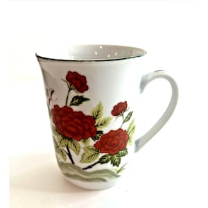 May include: White ceramic mug with a curved handle, featuring a floral design of red peonies and green leaves. The rim has a green trim. The mug is a standard size for coffee or tea.
