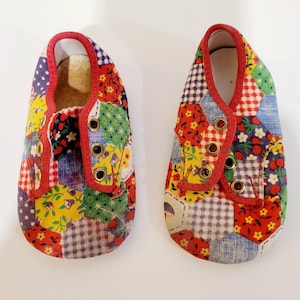 May include: A pair of baby shoes made of patchwork fabric. The shoes are red, yellow, green, blue, and white. They have a red trim and gold eyelets.