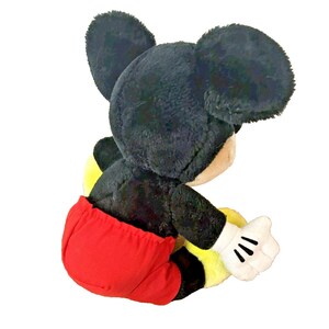 May include: A plush Mickey Mouse toy, featuring black fur, large round ears, and a red pair of shorts. The toy has white gloves and a yellow and black body. The toy is designed to sit.
