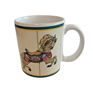 May include: Cream-coloured ceramic mug with a carousel horse design. The horse is tan with a saddle and decorative elements in red, gold, and blue. The mug has a green and yellow border and a white handle.