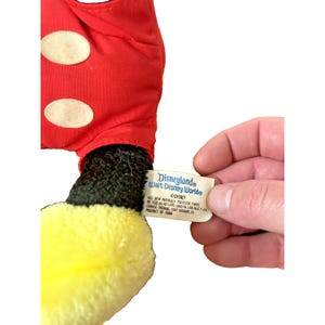 May include: A vintage Mickey Mouse plush toy, featuring a red outfit with white polka dots and yellow gloves. The tag reads "Disneyland Walt Disney Worlde". The plush is made of soft materials.