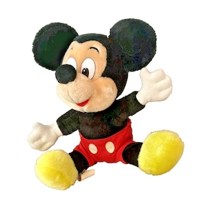 May include: A plush Mickey Mouse toy, featuring black fur, a tan face, and large black ears. Mickey wears red shorts with white buttons, white gloves, and yellow feet. The toy is posed with arms outstretched, and is sitting down.