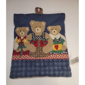 May include: A quilted pot holder featuring three teddy bears. The bears are appliquéd with details like aprons, flowers, and a basket of apples. The pot holder has a blue denim background with a plaid pattern at the bottom and a hanging loop.