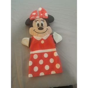 May include: A Minnie Mouse hand puppet. The puppet has a red dress with white polka dots, a white collar, and a red bow with white polka dots. The puppet has black ears and a smiling face.