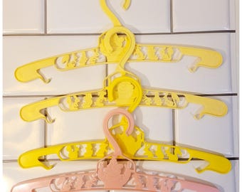 May include: Three plastic clothes hangers in yellow and pink. The hangers have a decorative design with cut-out shapes of cartoon characters. The hangers are designed for children's clothing and are hanging against a white tiled wall.