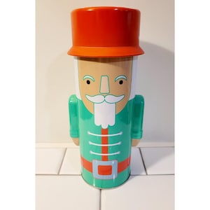 May include: A decorative tin shaped like a nutcracker soldier. The tin features a red hat, a green uniform with white accents, and a tan face with a white mustache and beard. The tin is designed with a festive holiday theme.