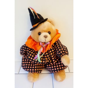 May include: A teddy bear in a Halloween costume. The bear is dressed in a black witch hat with orange trim and a matching polka-dot outfit. The outfit includes a ruffled orange collar and a bow with colourful ribbons.