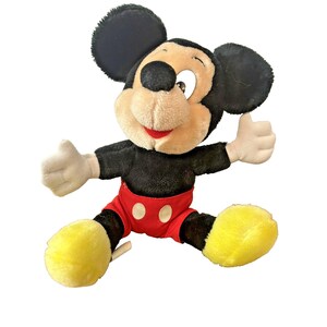 May include: A plush Mickey Mouse toy with black ears, a black body, red shorts with white buttons, and yellow feet. Mickey has a tan face, a red smile, and white gloves. He is giving a thumbs-up.