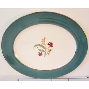 May include: An oval ceramic serving platter with a cream-coloured centre and a wide teal border. The centre features a delicate floral motif with small red flowers, green leaves, and yellow wheat-like stalks. The rim is finished with a gold trim.