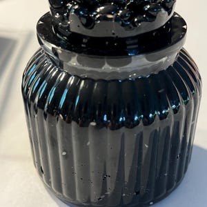 May include: A small, black, glass jar with a lid. The jar has vertical ribbed detailing and a rounded shape. The lid is adorned with a cluster of small, dark, rounded shapes. The jar is sitting on a white surface.