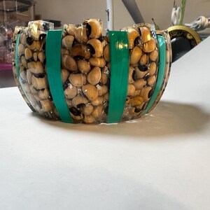 May include: A round bowl made of clear resin embedded with numerous tan black-eyed peas and accented with vertical, opaque teal green stripes. The bowl has a textured, uneven top rim and a transparent finish that reveals the organic bean shapes.
