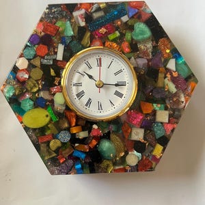 Pode incluir: Relógio hexagonal com moldura dourada e mostrador branco com algarismos romanos pretos. O relógio está inserido num mosaico de peças de vidro coloridas e de formato irregular.