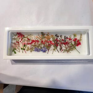 Puede incluir: Una pieza rectangular de resina blanca con una colección de flores secas de varios colores. Las flores incluyen flores rojas, rosas, moradas y amarillas, dispuestas horizontalmente. La resina transparente permite una visibilidad total de las flores.
