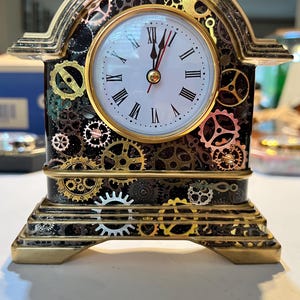 May include: A decorative clock with a steampunk design. The clock features a white face with black Roman numerals and gold hands. The clock's frame is black with gold and copper-colored gears.
