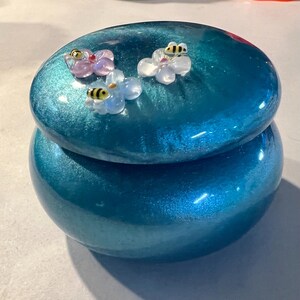 May include: A teal trinket box with a glossy finish. The lid is decorated with three glass flowers, each with a small bee. The box is designed for small items and adds a touch of whimsy. The box is approximately 10 cm in diameter.