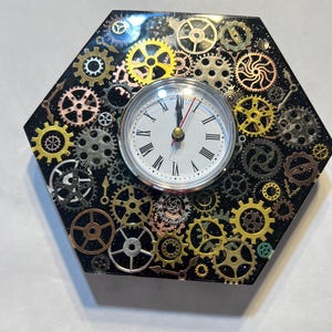 May include: Hexagonal clock with a black background, adorned with various metallic gears in gold, silver, and copper tones. The clock face is white with black Roman numerals and red hands.