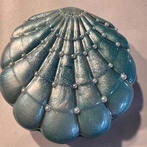 May include: A pearlescent blue and silver seashell-shaped trinket box. The box is adorned with small, white pearl accents along the edges. The shell design has a textured, fan-like pattern.