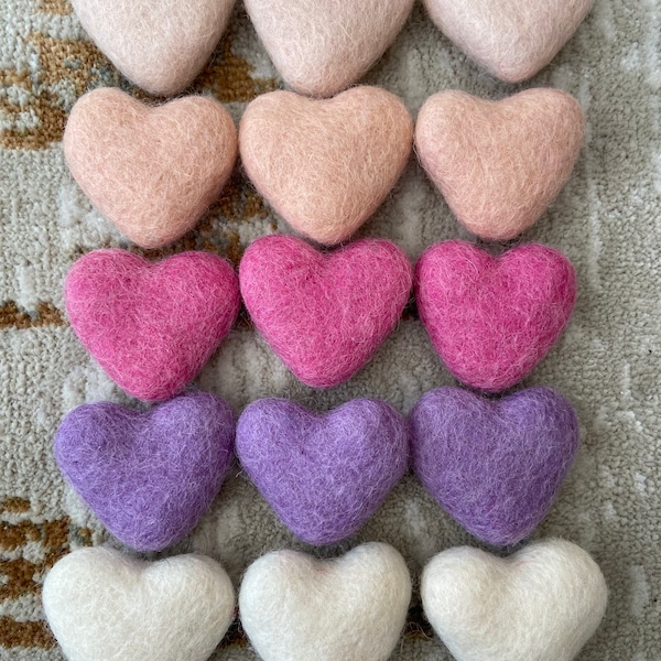 Peut inclure: Un ensemble de 18 décorations en forme de cœur en feutre dans différentes nuances de rose, violet et blanc. Les cœurs sont disposés en trois rangées de six, chaque rangée présentant une couleur différente.