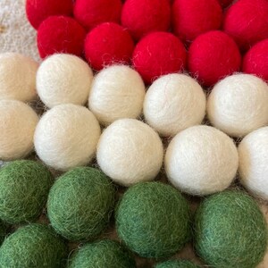 May include: A close-up of a felt ball garland with red, white, and green felt balls. The garland is arranged in a triangular shape, with the red balls at the top, the white balls in the middle, and the green balls at the bottom.