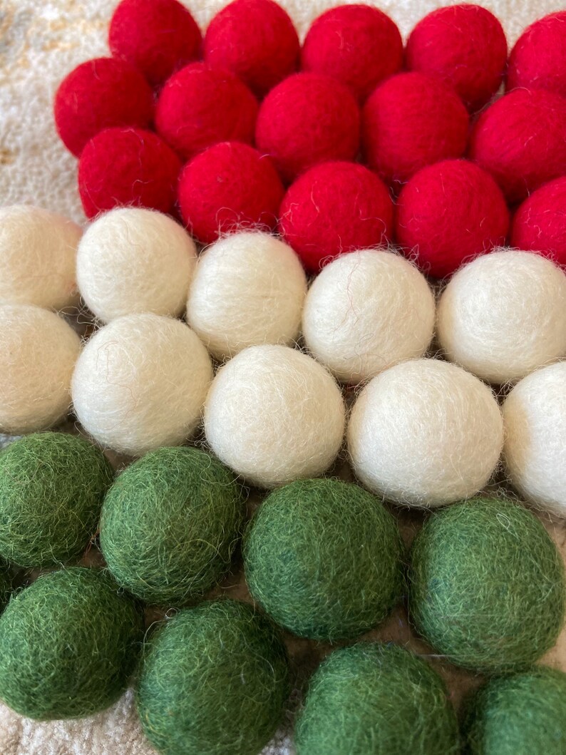 May include: A close-up of a felt ball garland with red, white, and green felt balls. The garland is arranged in a triangular shape, with the red balls at the top, the white balls in the middle, and the green balls at the bottom.