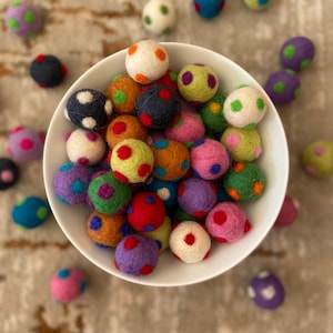 Può includere: Una ciotola bianca piena di palline di feltro colorate. Le palline sono decorate con pois bianchi e hanno una varietà di colori, tra cui rosso, verde, blu, giallo, viola e arancione.