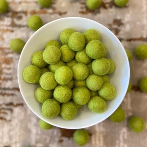 May include: A white bowl filled with approximately 30 small, green felt balls. The bowl is sitting on a patterned rug. The text '#13 Avocado' is visible below the bowl.