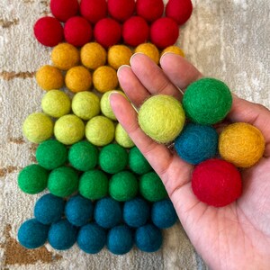 Puede incluir: Un surtido de bolas de fieltro arcoíris en colores rojo, naranja, amarillo, verde, azul y turquesa. Las bolas están dispuestas en forma de pirámide, con la bola más grande en la parte inferior y la más pequeña en la parte superior.