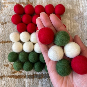 May include: A hand holding a variety of felt balls in red, white, and green colors. The balls are arranged in a pattern on a surface.