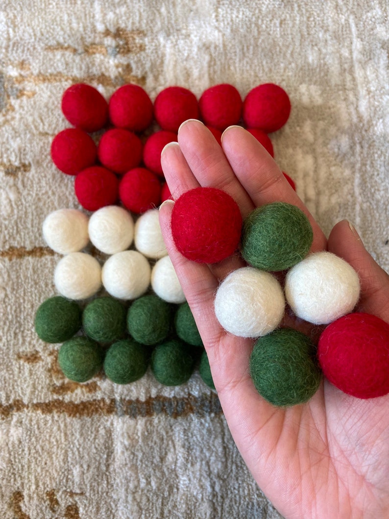 May include: A hand holding a variety of felt balls in red, white, and green colors. The balls are arranged in a pattern on a surface.