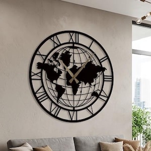 May include: A large circular wall clock featuring a black metal frame with Roman numerals and a central world map silhouette. The clock has thin gold-colored hands and is mounted on a light-colored wall above a gray sofa in a living room setting.