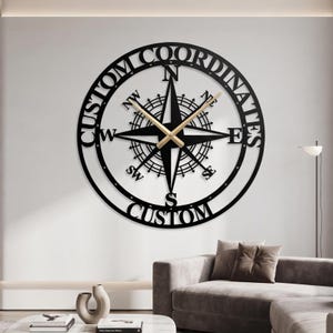 May include: A circular wall clock featuring a black metal compass rose design with the text CUSTOM COORDINATES and CUSTOM integrated into the frame. The clock has gold-colored hour and minute hands centered on the compass dial with cardinal directions.