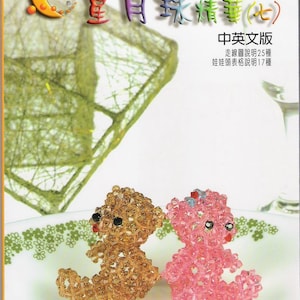 May include: Two beaded teddy bears, one brown and one pink, are sitting on a white plate with a green floral pattern. The bears are made of small, round beads and have black eyes and red mouths. The book cover in the background has the title "Star Moon Bead Essence (7)" in Chinese and English.