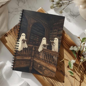 May include: Spiral-bound notebook featuring a whimsical illustration of three ghost figures reading books in a vintage library setting. The artwork showcases a detailed wooden staircase and bookshelves, with the ghosts in flowing white sheets. The notebook has a dark, gothic aesthetic.